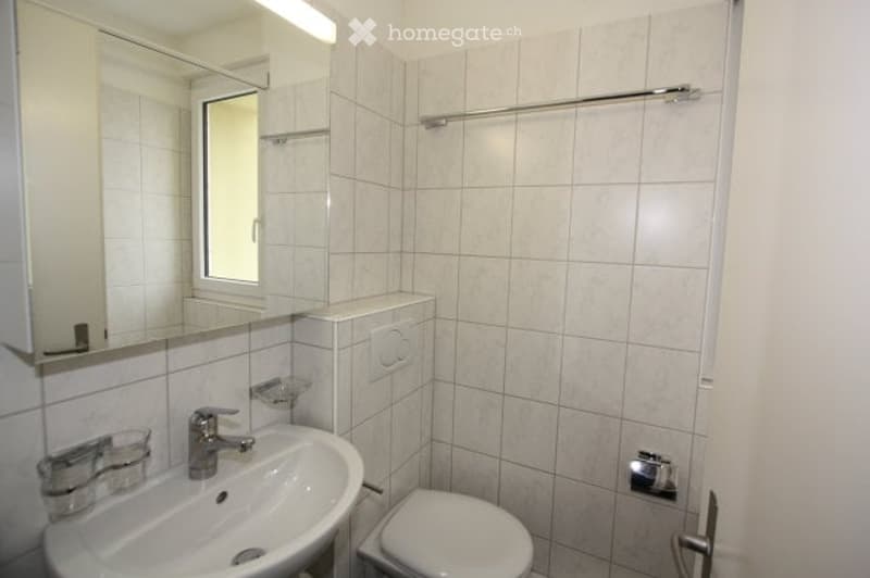 A small, clean bathroom with white tiled walls and floor, a sink with a mirror above it, a toilet, and a towel rack on the wall.