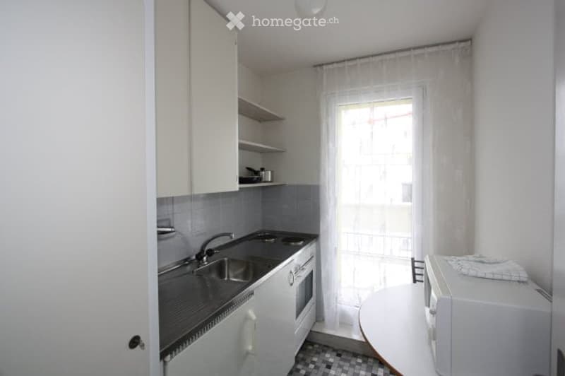 Modern kitchen with white cabinets, stainless steel sink, oven, microwave, and a small table with a chair. The space features a tiled floor and a window with sheer curtains.