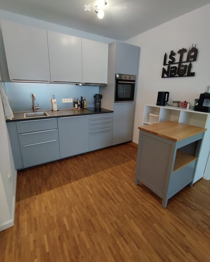 Modern kitchen with light grey cabinets, wooden flooring, and a central island. Features include a sink, faucet, oven, and various kitchen appliances.