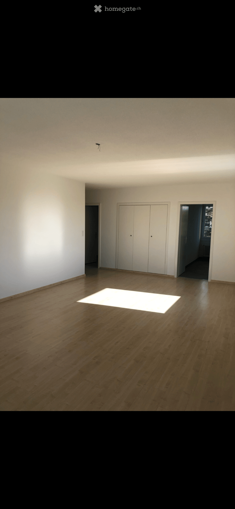 Empty room with wooden flooring, white walls, and a large illuminated square in the center. There are two white closet doors and an open doorway leading to another space.