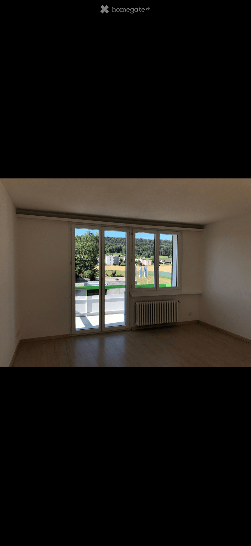 Empty room with wooden flooring, large sliding glass doors leading to a balcony, radiator, and a view of greenery and buildings.