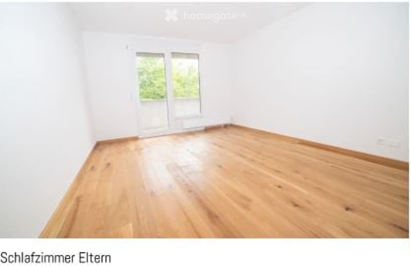 Empty room with light wooden flooring and white walls, featuring a large window with a view of greenery outside.