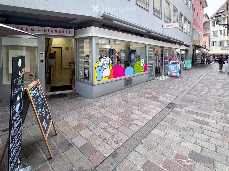 A storefront with a glass display window showing colorful graphics and text, with a sidewalk and other shops in the background.