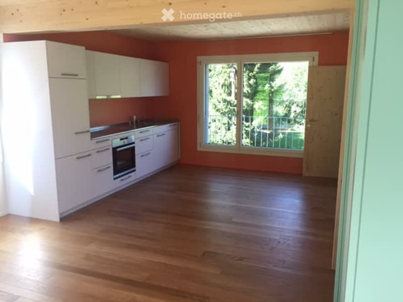 Bright kitchen with white cabinets, a built-in oven, and a large window overlooking a green outdoor area. The floor is made of polished wood.