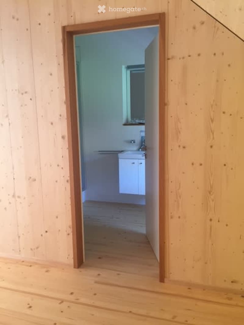 An unfurnished room with light wooden walls and flooring, featuring an open doorway leading to a small, white-walled bathroom with a sink and mirror.