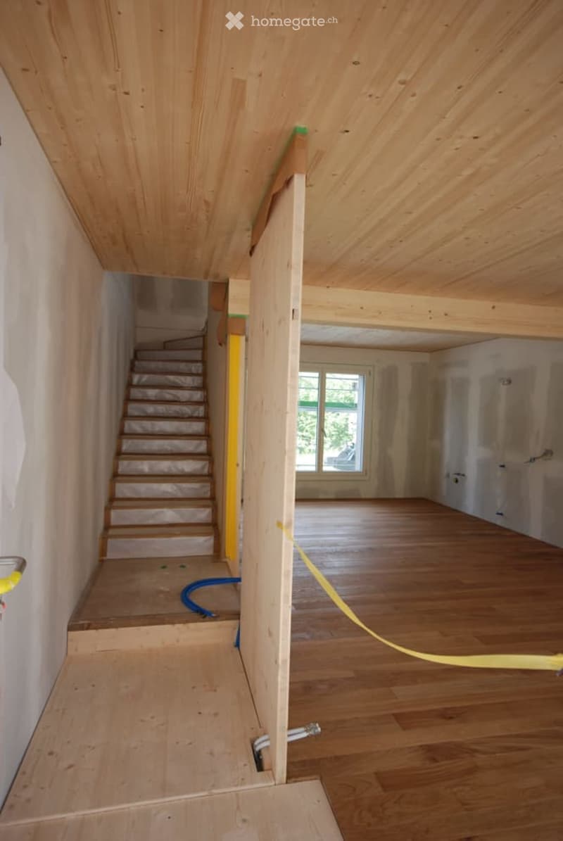 Empty room with wooden flooring and exposed wooden ceiling beams. Stairs lead to another level, and a window is visible in the background.