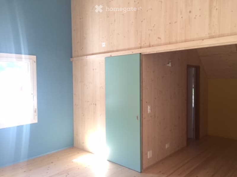 Empty room with wooden flooring, blue walls, and a large window allowing natural light to enter. The room features a sliding door and exposed wooden beams on the ceiling.