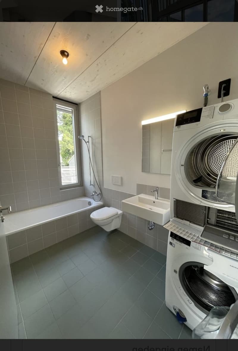 Modern bathroom with a bathtub, toilet, sink, and washing machine. Features tiled walls, a window, and a sleek design.