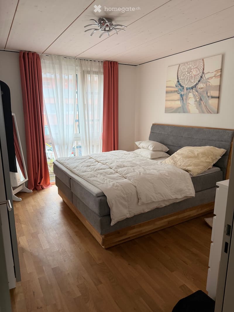 Cozy bedroom with a large bed, pillows, and a grey headboard. The room features wooden flooring, sheer white curtains, and a large picture frame on the wall.