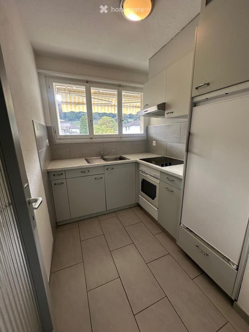 Modern kitchen with light grey cabinets, white countertops, a sink, oven, refrigerator, and a range hood. The room features tiled flooring and large windows with yellow blinds.
