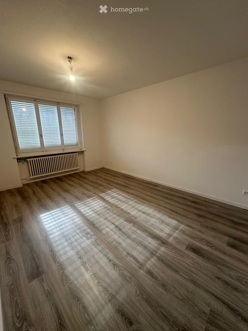 Empty room with wooden flooring, white walls, large window with blinds, radiator, and ceiling light fixture.