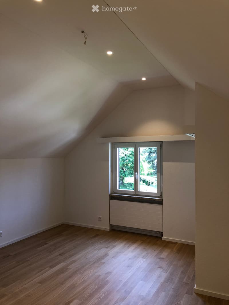 Empty attic room with wooden flooring, white walls, and a large window offering a view of greenery outside. The room features a radiator below the window and recessed ceiling lights.