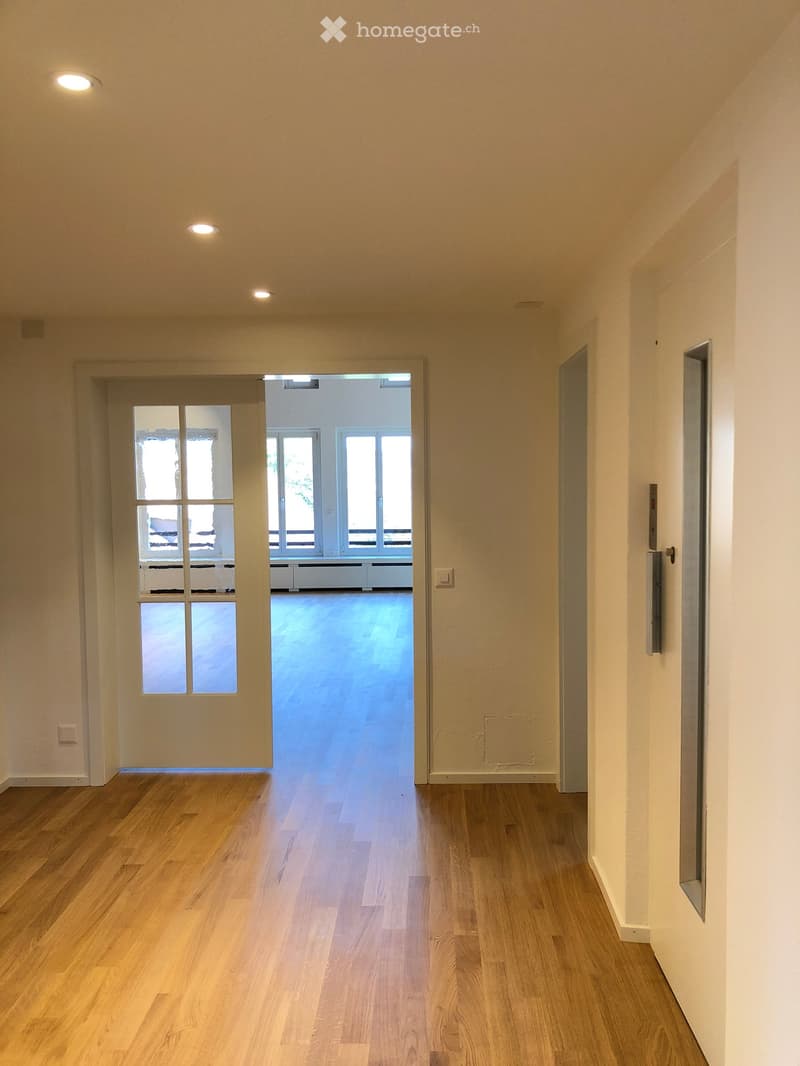 Empty hallway with wooden flooring, white walls, recessed lighting, and a door leading to a brightly lit room with large windows.