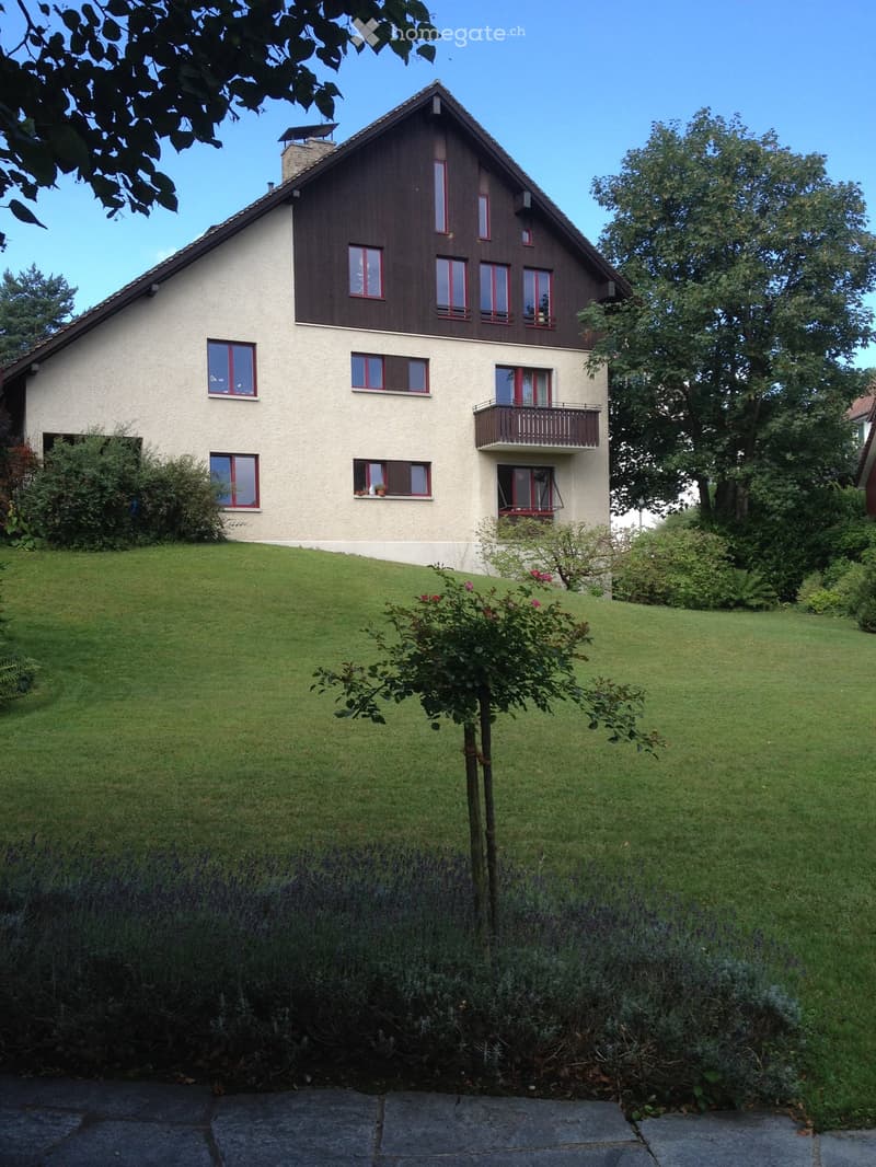 Two-story house with a sloped roof, beige walls, red-framed windows, and a balcony on the upper floor. The house is surrounded by a well-maintained lawn and garden with various plants and trees.