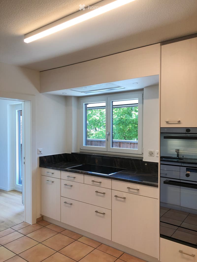 Modern kitchen with white cabinets, black countertop, and stainless steel appliances, including a sink and oven. Features large windows and a skylight.
