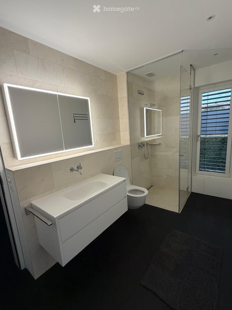 Modern bathroom with a white sink, toilet, large mirror, and glass-enclosed shower. Light-colored tiles and black flooring.
