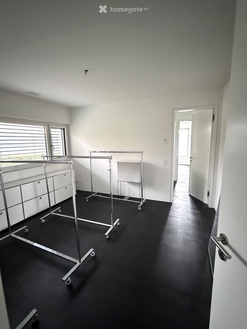 Empty room with black flooring, white walls, large windows with blinds, and several clothing racks.