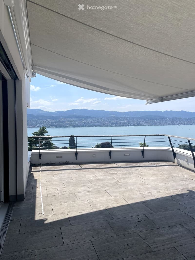 Modern balcony with wooden decking, metal railing, and retractable awning, overlooking a scenic lake and distant mountains.