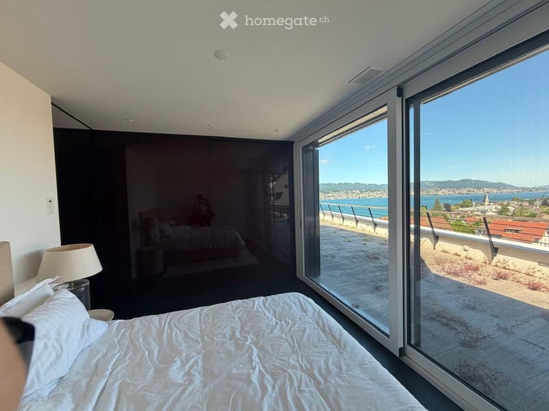 Modern bedroom with large windows offering a scenic ocean view, featuring a neatly made bed and a bedside lamp.