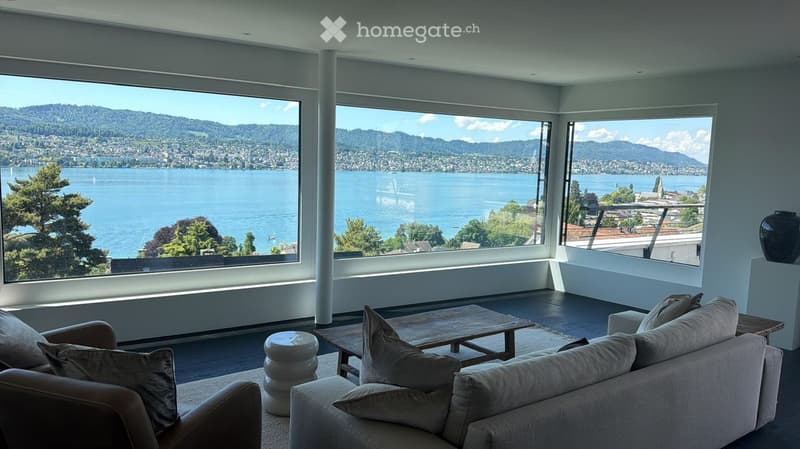Modern living room with large windows offering a scenic lake and mountain view, featuring a sofa, coffee table, and decorative stool.