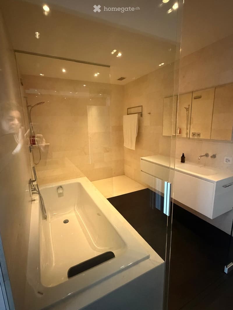 Modern bathroom with a large bathtub, glass-enclosed shower, white sink, and mirror. Features a sleek black floor and warm lighting.
