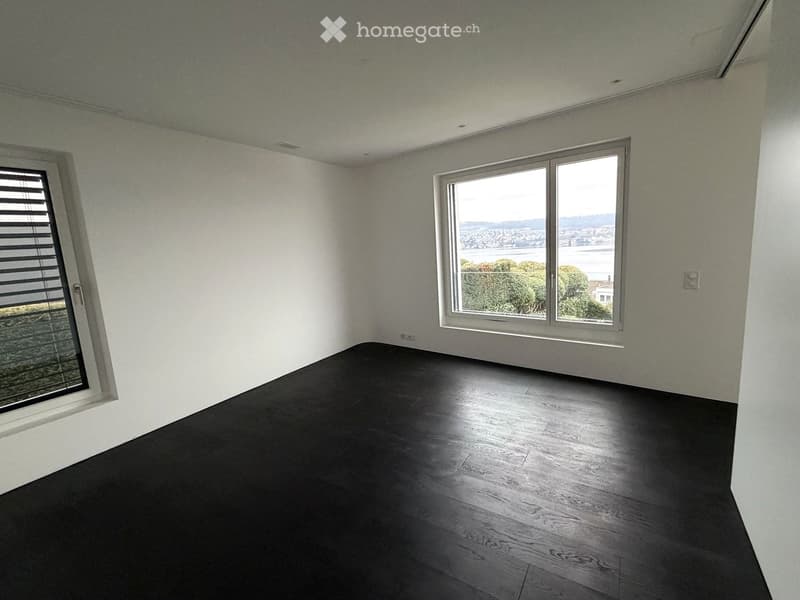 Empty room with dark wooden flooring, white walls, large windows offering a scenic view, and a sliding door with blinds.