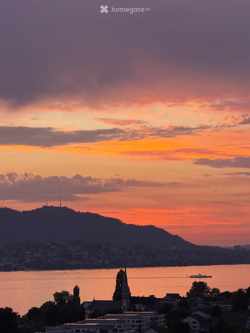 A panoramic view of a cityscape at sunset, featuring a lake, a church spire, and distant hills.