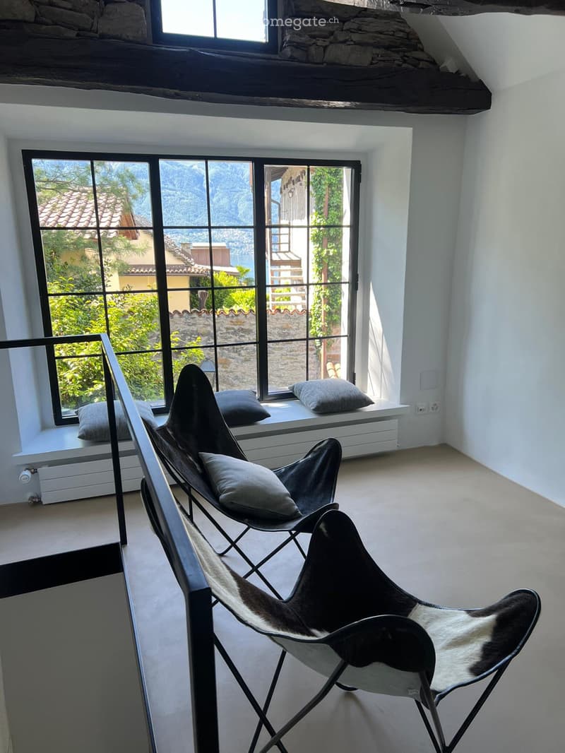 Modern furnished room with large windows offering a scenic view, featuring sleek black chairs with fur cushions and a window seat with additional cushions.
