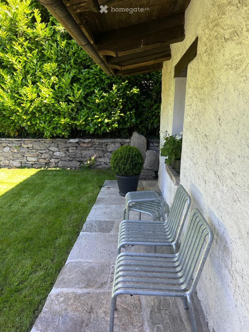 Outdoor patio area with stone flooring, metal chairs, small potted plant, and a stone wall with greenery in the background.