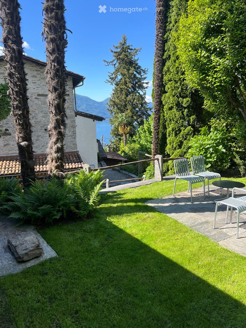 A well-maintained garden with a lawn, a stone pathway, and outdoor furniture including chairs and a table, surrounded by tall trees and a view of a body of water and mountains in the background.