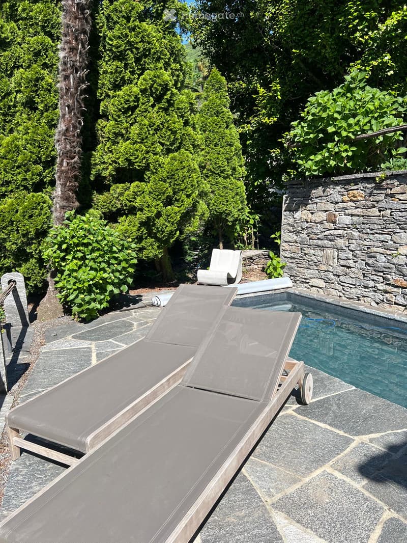 Outdoor patio with two sun loungers, a small swimming pool, and a stone wall, surrounded by lush greenery and trees.