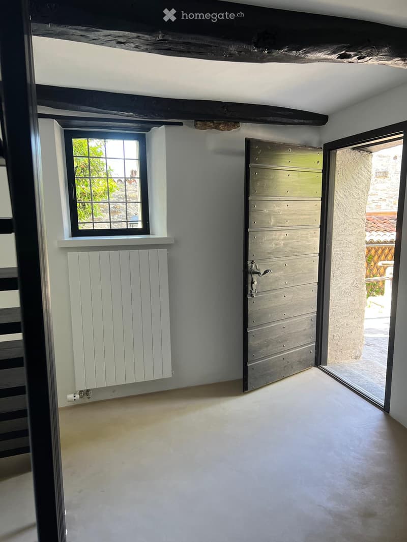 Empty room with white walls, wooden ceiling beams, a window with a radiator below, and a closed wooden door leading outside.