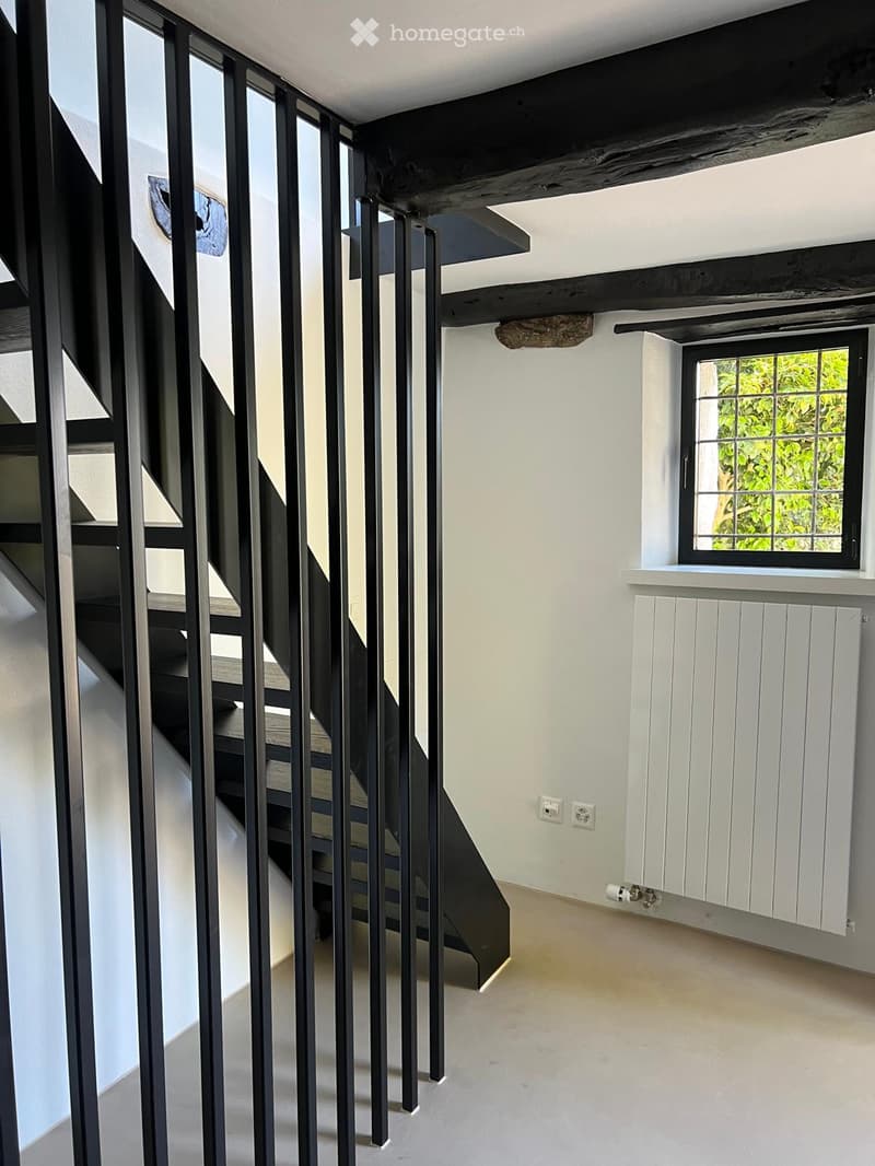 Empty room with white walls, a staircase with black metal railings, a window with a view of greenery, and a white radiator on the wall.