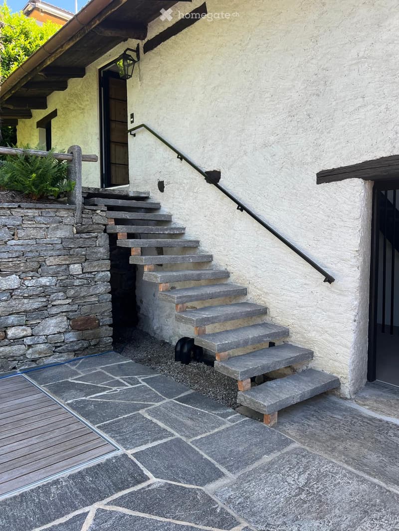 Stone steps leading up to a house entrance, with a metal railing and a small garden area on the left.