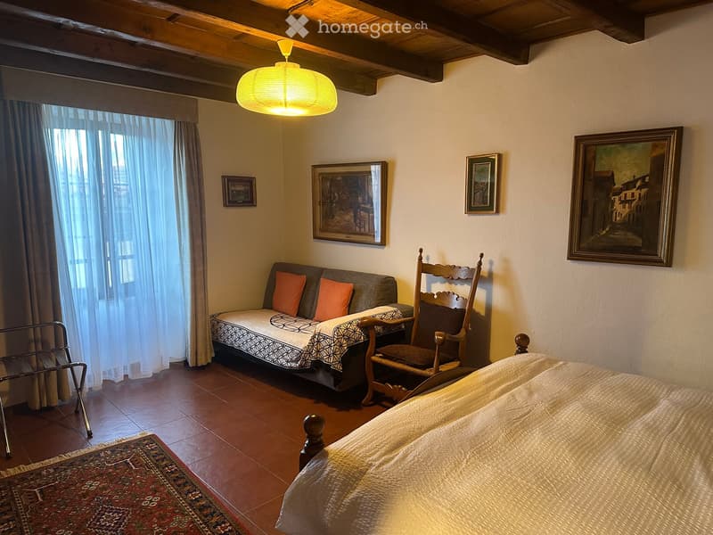 Cozy bedroom with a bed, sofa, wooden chair, and decorative artwork on the walls. Features exposed wooden beams and warm lighting.