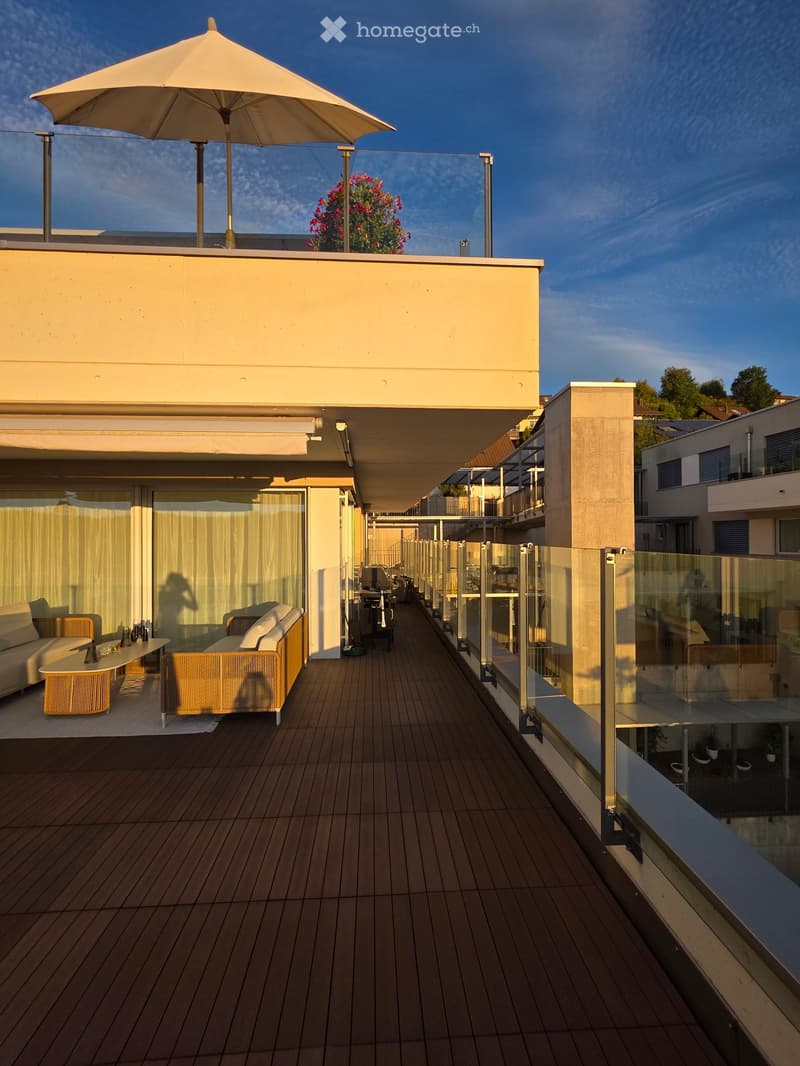 Modern rooftop terrace with wooden decking, glass railing, and outdoor furniture including a sofa, table, and umbrella.