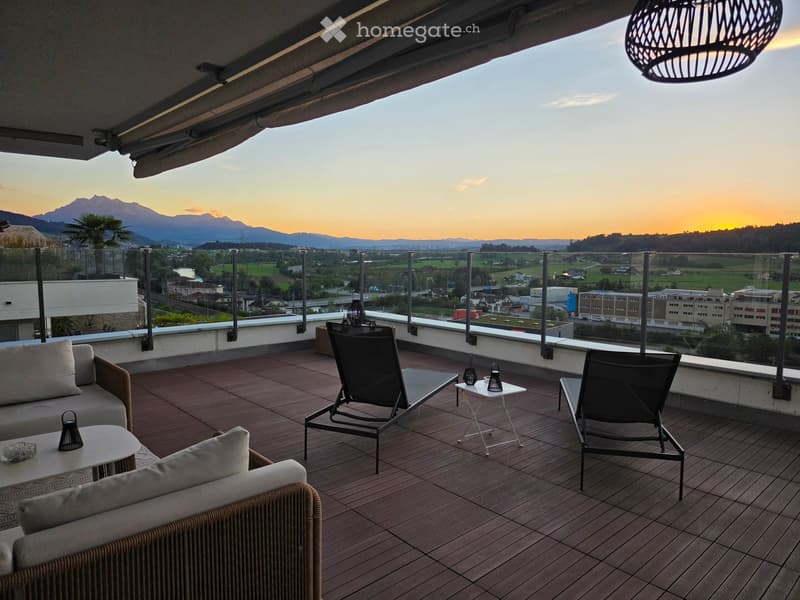 Balcony with wooden decking, sofa, chairs, small tables, and a scenic mountain view at sunset.