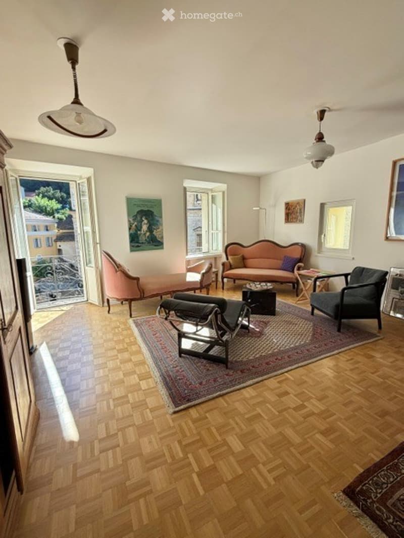 Living room with wooden flooring, large windows, a view, a sofa, armchairs, coffee table, ottoman, floor lamp, and mirrors.