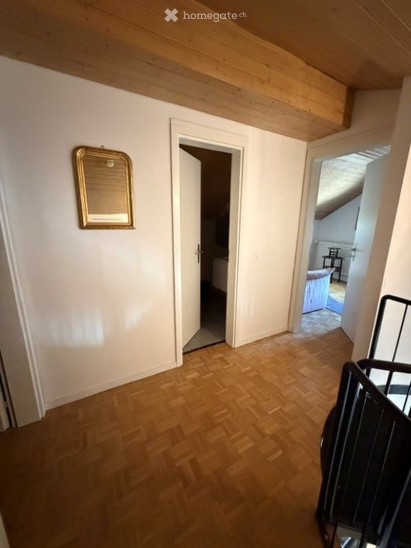 Empty room with wooden flooring, white walls, and a slanted wooden ceiling. A mirror is mounted on the wall, and there are open doorways leading to other rooms.