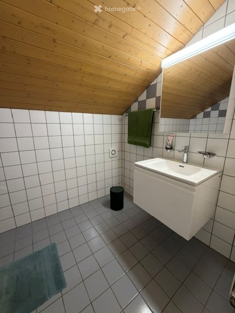 Modern bathroom with white tiles, wooden ceiling, white sink, mirror, and a green towel hanging on the wall.