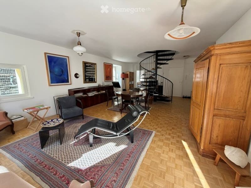 Spacious living room with wooden flooring, large window, modern furniture including a sofa, chairs, coffee table, ottoman, and a stylish floor lamp. A spiral staircase leads to an upper level, and the room is decorated with artwork and a bookshelf.