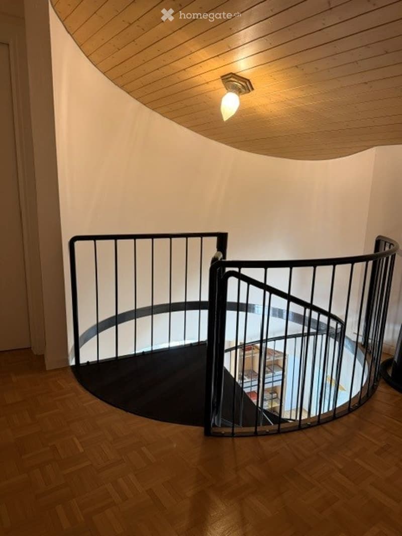 Spiral staircase with wooden floors, white walls, and a modern black metal railing. The ceiling is wooden with integrated lighting.