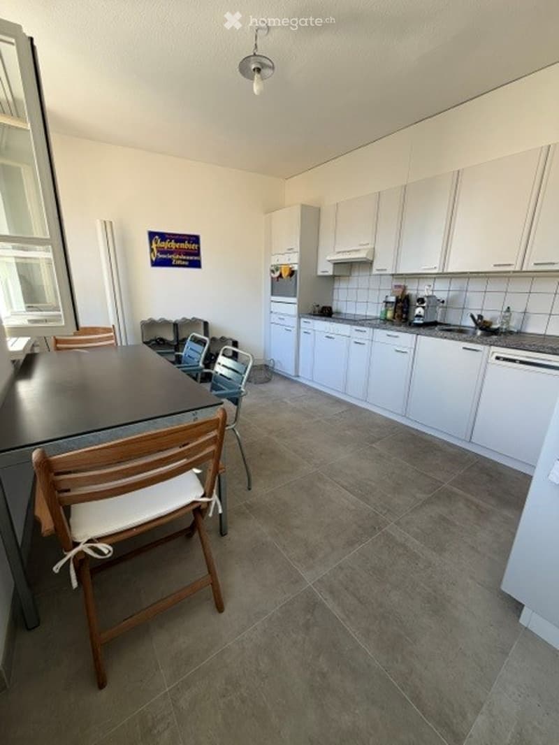 Modern kitchen with white cabinets, a central island, and a dining area with chairs and a table. The floor is tiled, and there is a sink with a faucet on the counter.