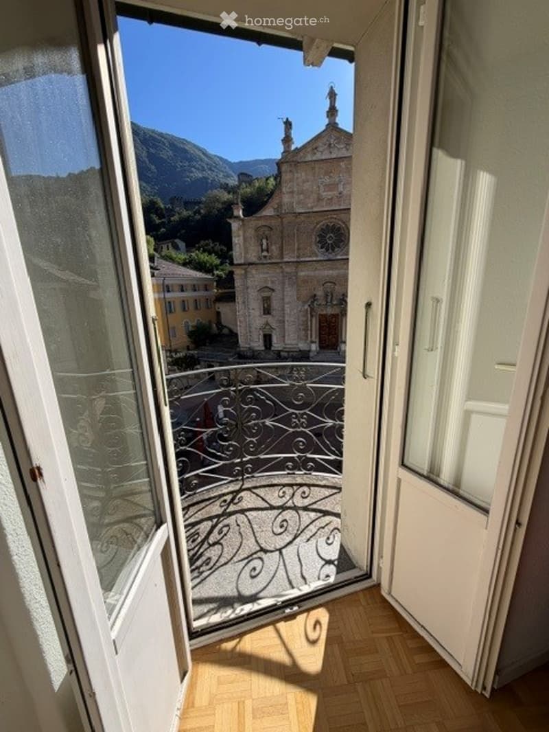Balcony with ornate metal railing, overlooking a town square with a church and mountains in the background.