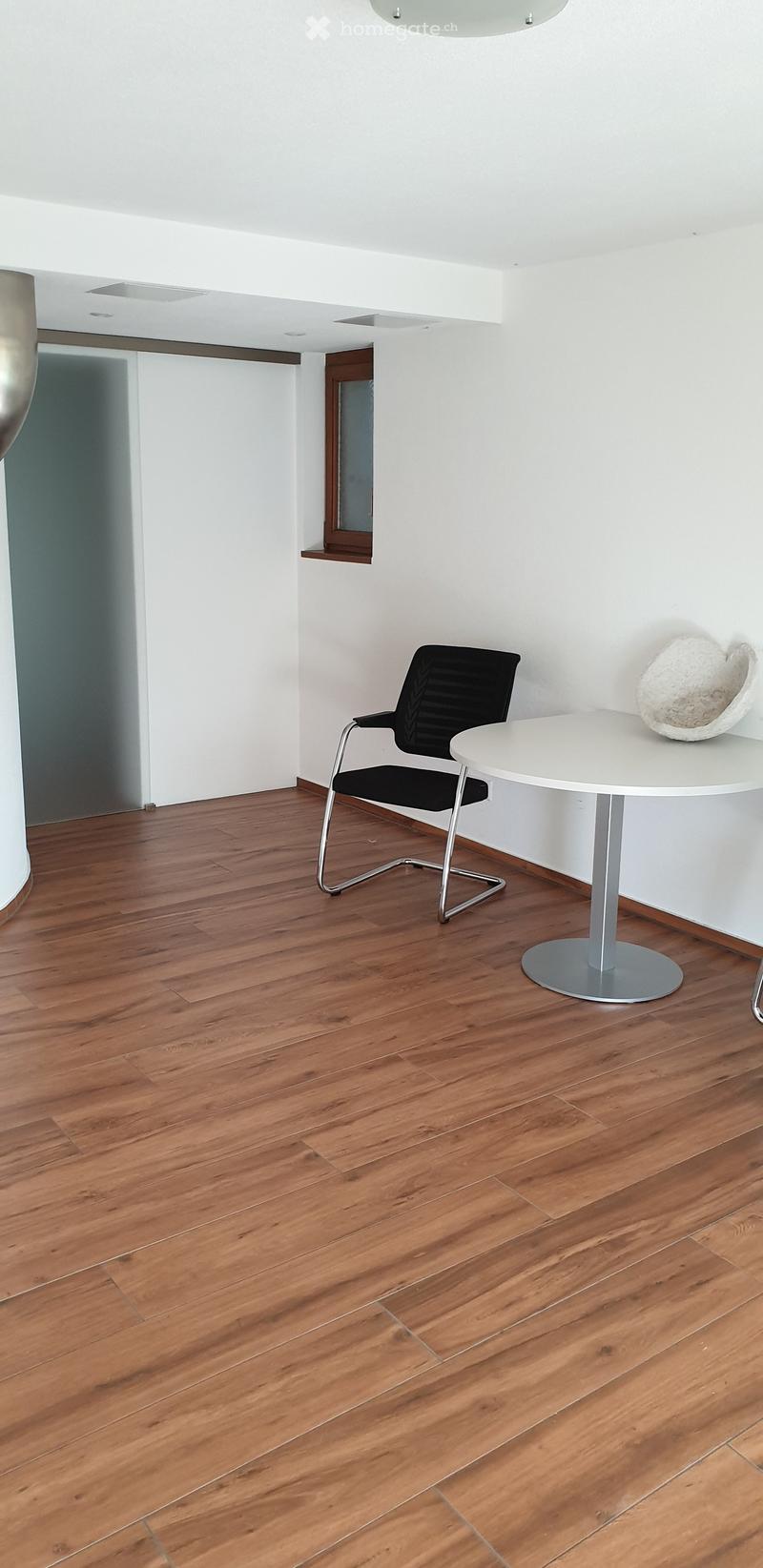Minimalist room with wooden flooring, white walls, a single black chair, a white round table, and a decorative sculpture on the table.