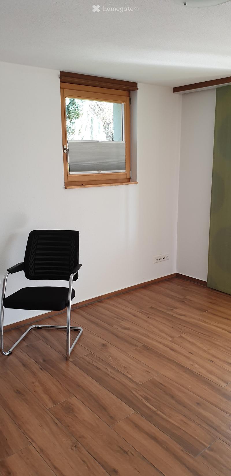 A small, unfurnished room with wooden flooring, white walls, and a single black office chair near a window with blinds.