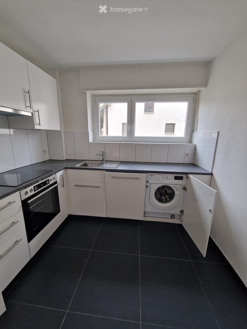 Modern kitchen with white cabinets, black countertops, built-in appliances including stove, oven, dishwasher, and washing machine, ceramic tile flooring, and a window overlooking a building.