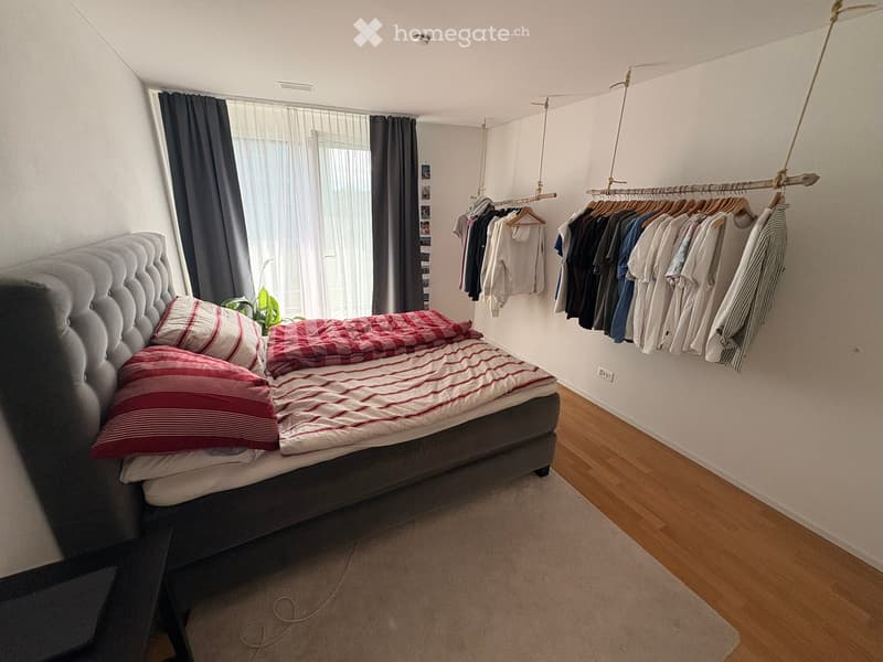 A modern bedroom with a tufted headboard bed, red and white striped bedding, a clothing rack with hanging shirts, and a large window with curtains.