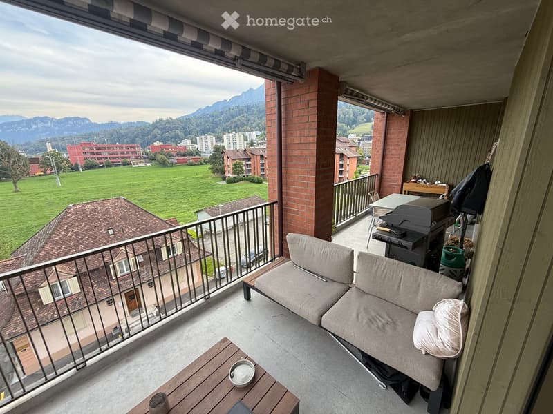 Balcony with a view of a green field and mountains, furnished with a sofa, small table, grill, and chairs.