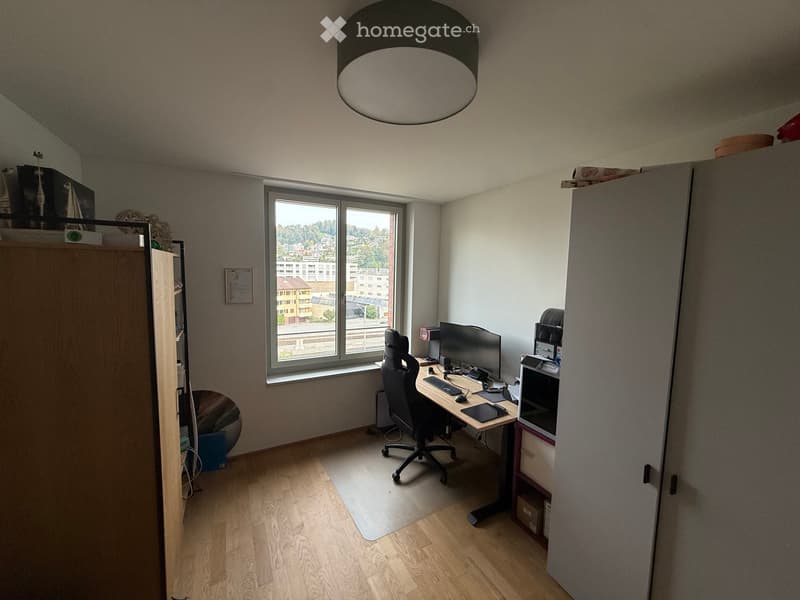 A modern office space with a wooden floor, large window offering a city view, a desk with a computer setup, a comfortable office chair, and built-in storage cabinets.